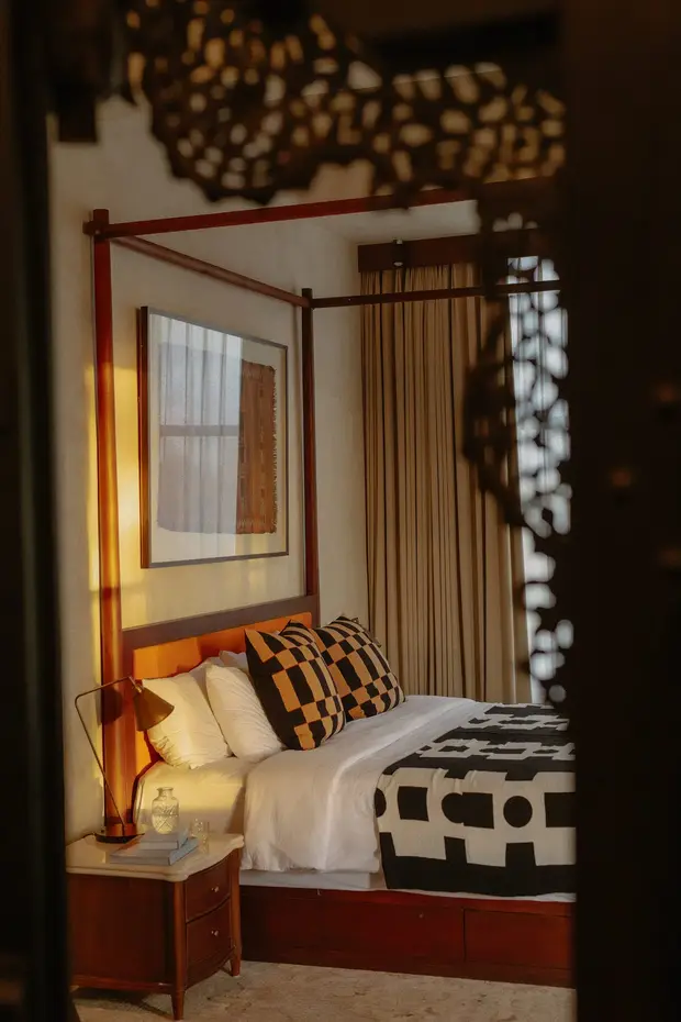 A timber four poster bed with a brass lamp and patterned cushions at Rumah Buaya